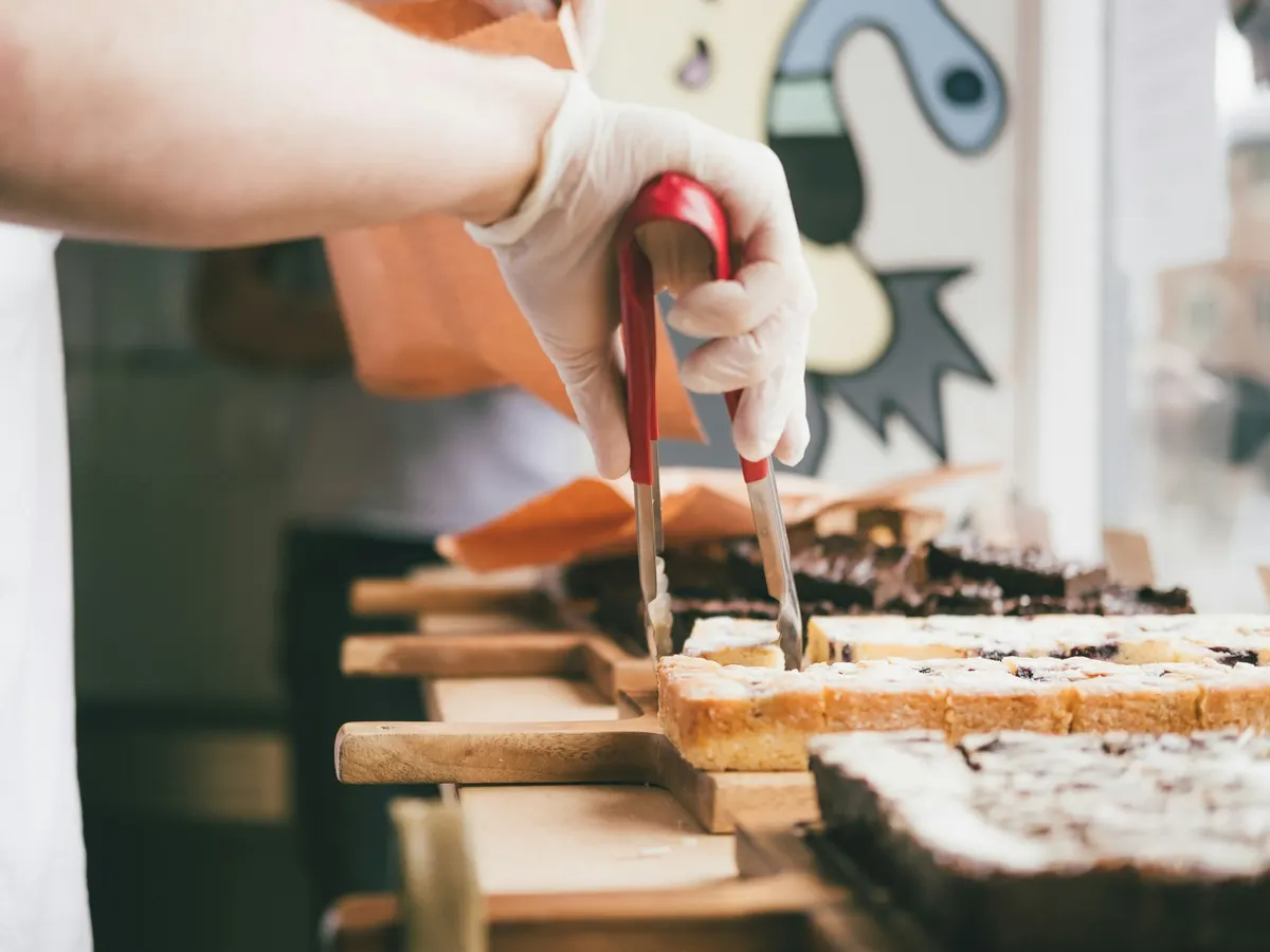 Mão com luva a servir bolos com uma pinça ao balcão de uma pastelaria